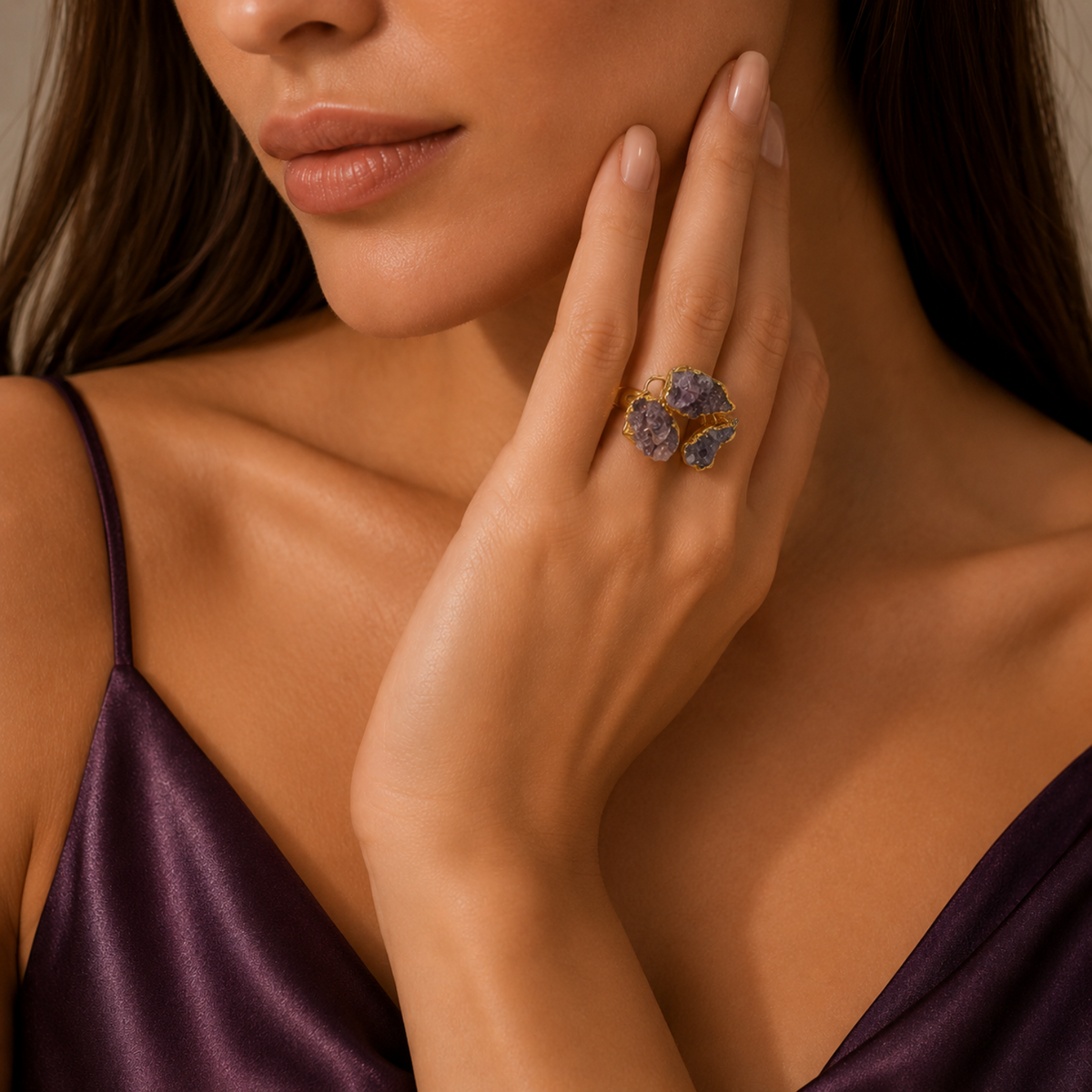 A close-up lifestyle shot of a model wearing a gold ring with purple and blue gemstones on her finger, wearing a dark purple satin top.