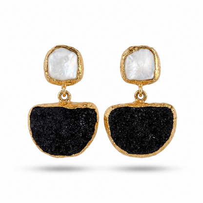 A pair of gold drop earrings featuring a textured black druzy stone in a large semicircle and a small irregular white pearl stud above, set in gold bezels on a white background.