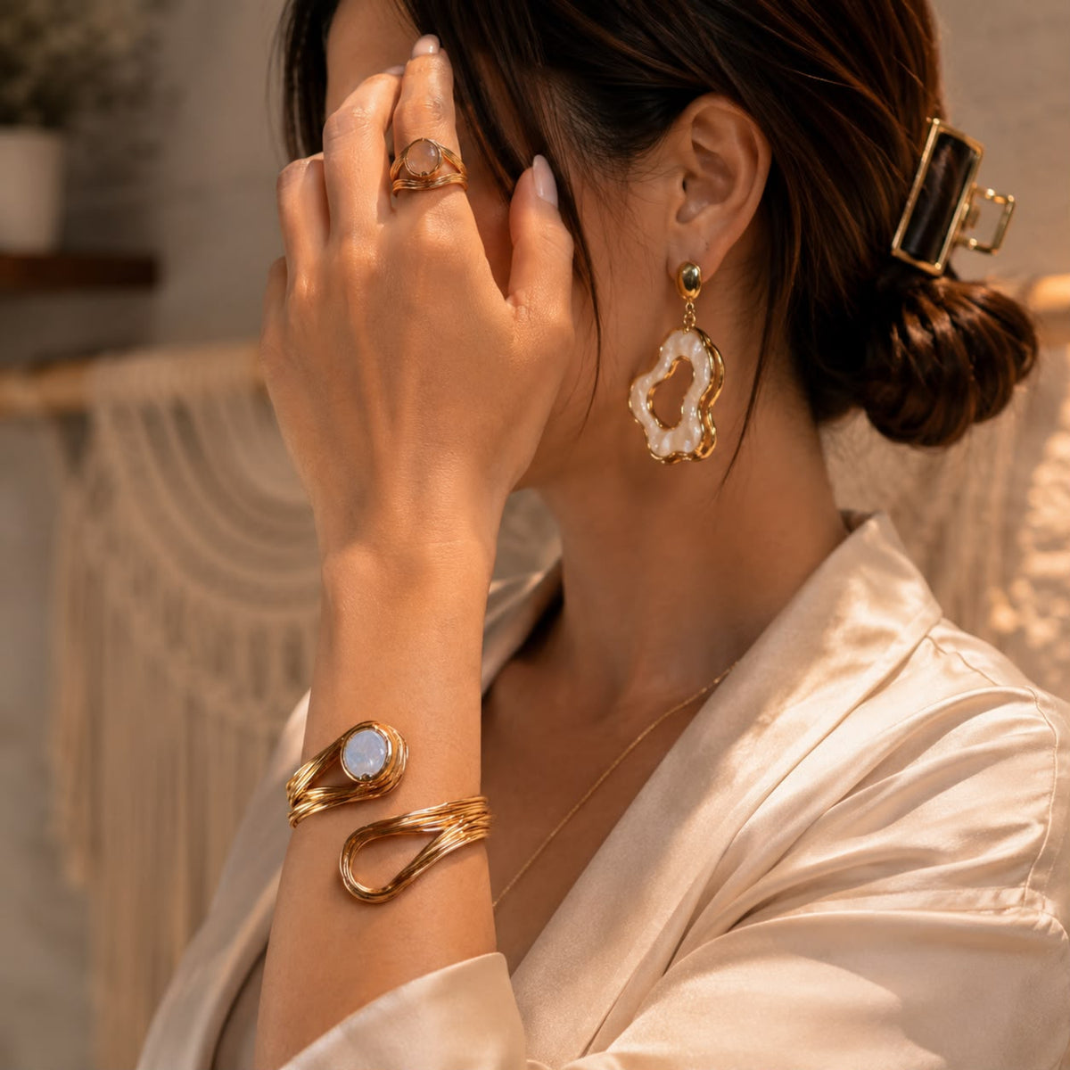 A woman wearing gold jewelry, including a gold wrap bracelet with a white stone, a gold ring, and a gold dangle earring with a white agate-like cutout, styled with a cream satin shirt in a warm indoor setting.