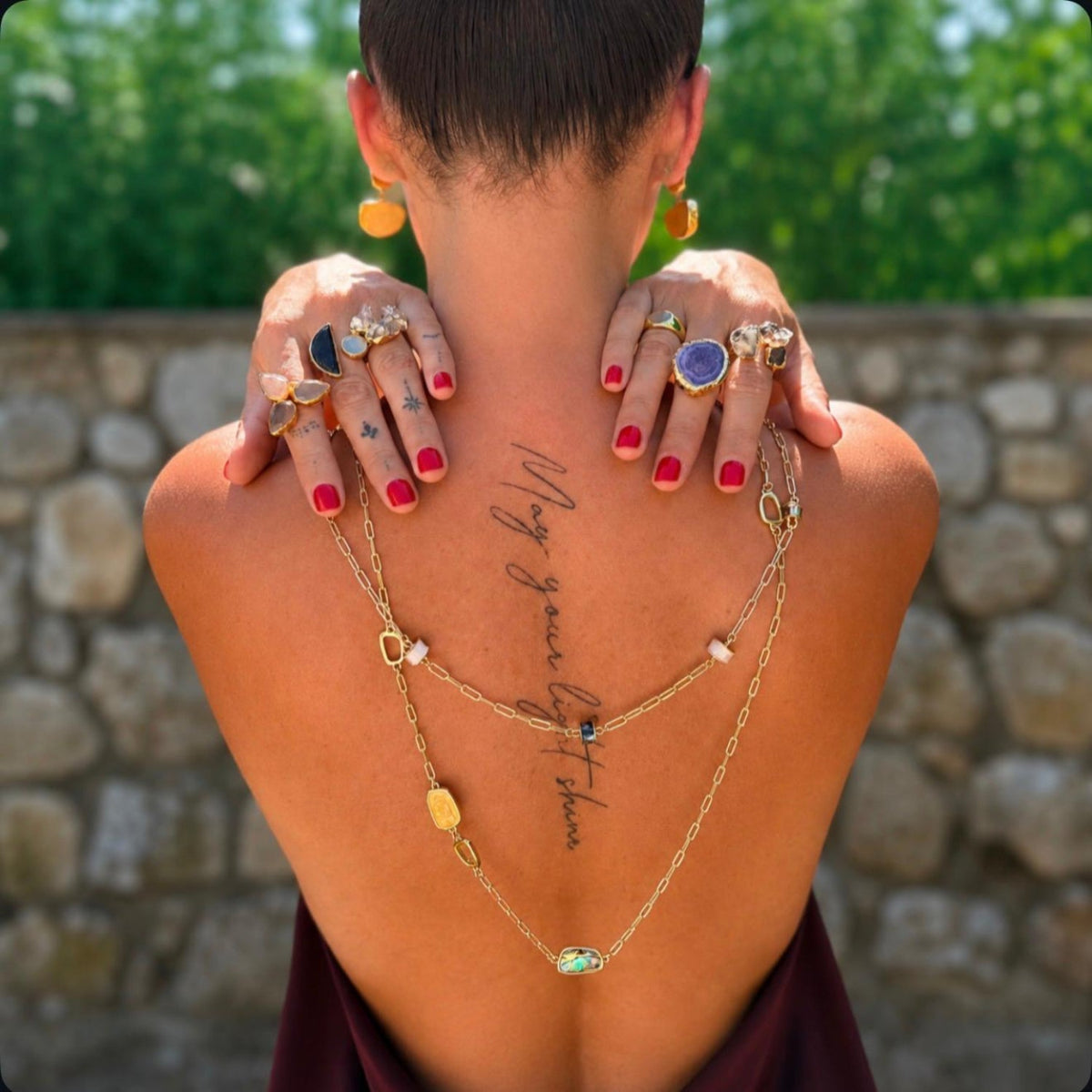 A woman viewed from behind outdoors, wearing a layered gold necklace with small stones and rings on her hands, with green foliage and a stone wall in the background.
