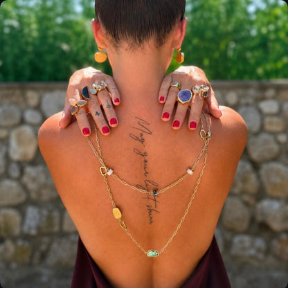 A woman viewed from behind outdoors, wearing a layered gold necklace with small stones and rings on her hands, with green foliage and a stone wall in the background.