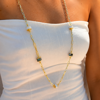 A gold chain necklace with small rectangular green and black marbled beads and a white disc accent, worn on a model's neck over a white strapless top.