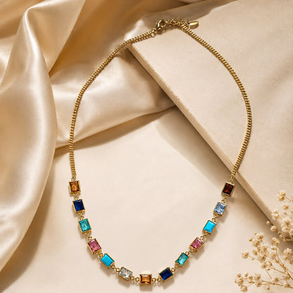 A gold chain necklace with a series of colorful rectangular gemstones in blue, pink, orange, and clear, styled on a beige background with draped satin fabric and small dried flowers.