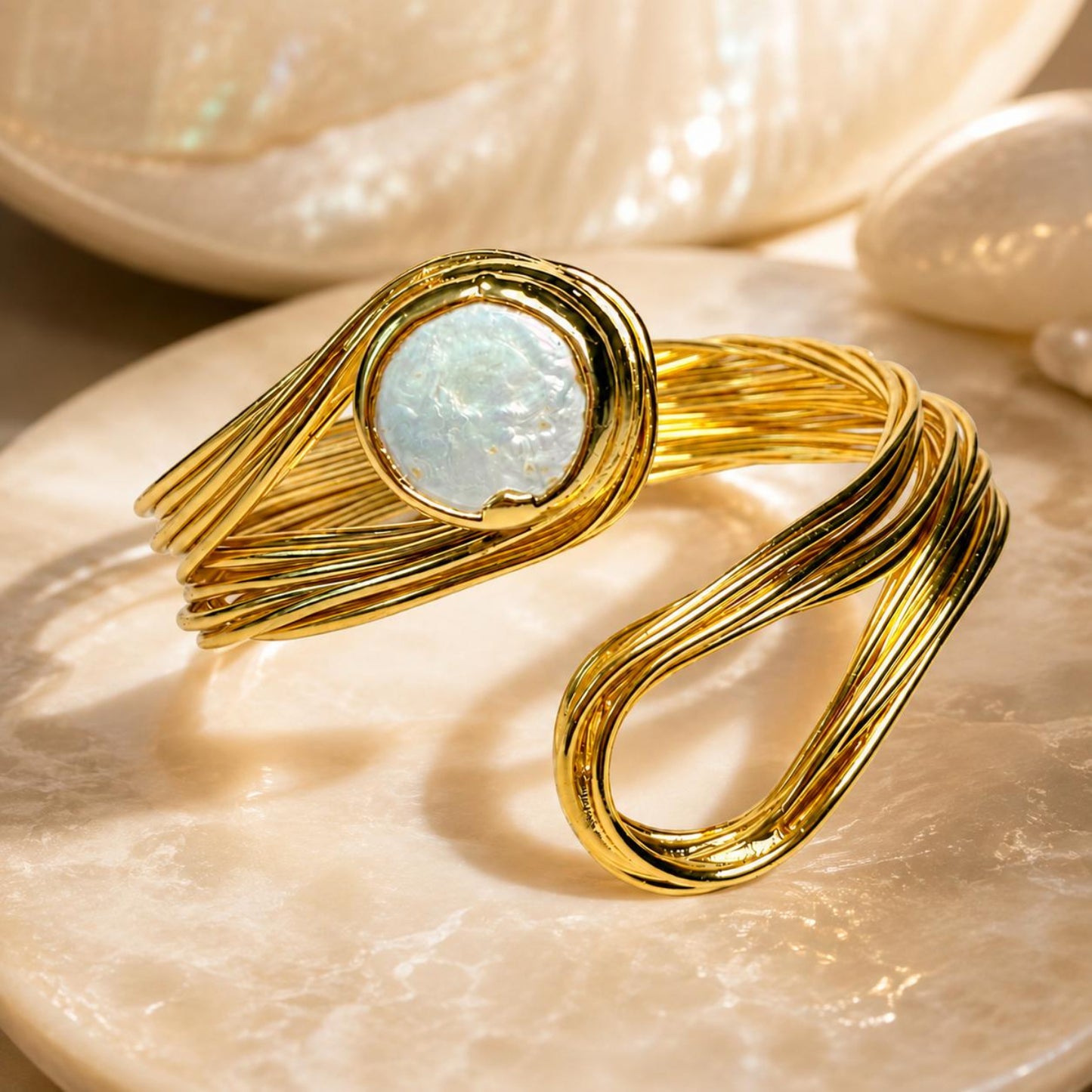 A gold-toned wrap cuff bracelet with a round iridescent white stone, displayed on a polished stone surface with shell props in the background.