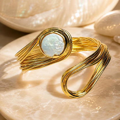 A gold-toned wrap cuff bracelet with a round iridescent white stone, displayed on a polished stone surface with shell props in the background.