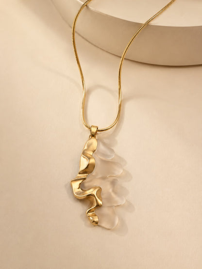 A gold chain necklace with an abstract, wavy gold pendant layered over a translucent, layered white shell or glass piece, displayed on a warm beige background.