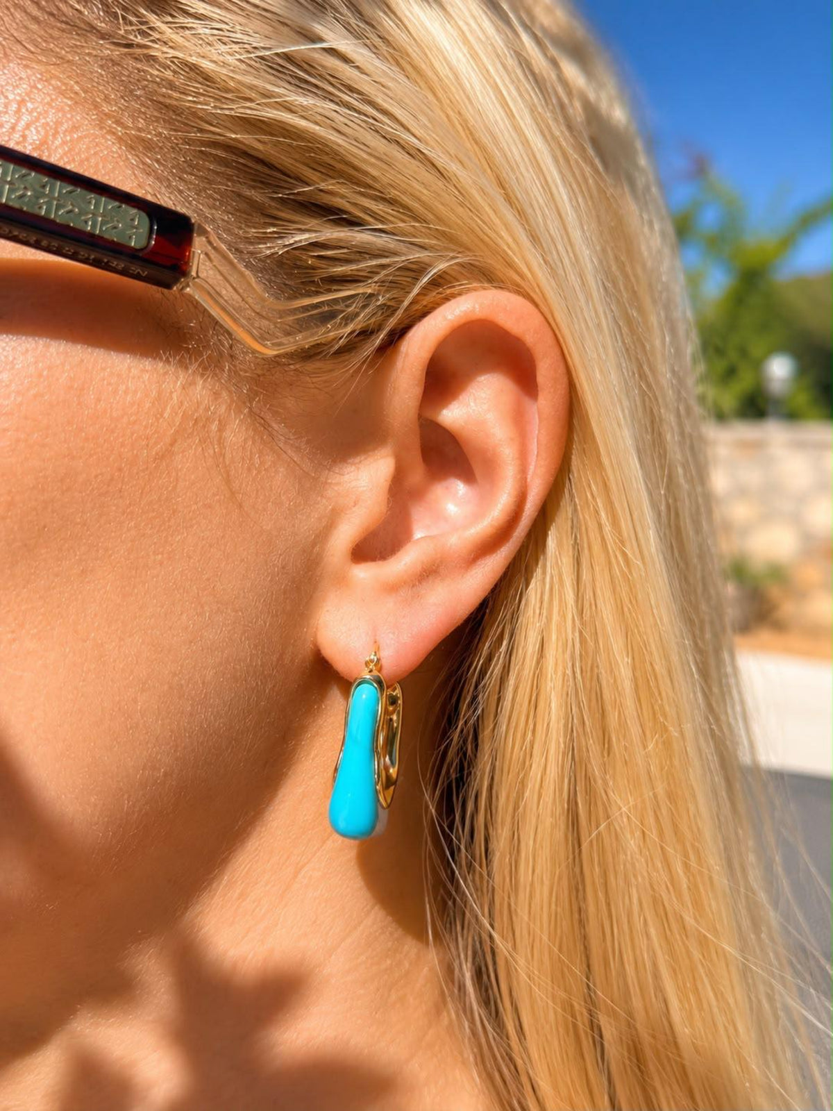 A close-up of a woman's ear wearing a turquoise drop earring with gold hoop earrings, outdoors under bright sunlight.