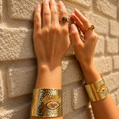 A close-up of a person's hands and forearms resting against a light beige brick wall, wearing two wide textured gold cuff bracelets with an eye motif and two rings.