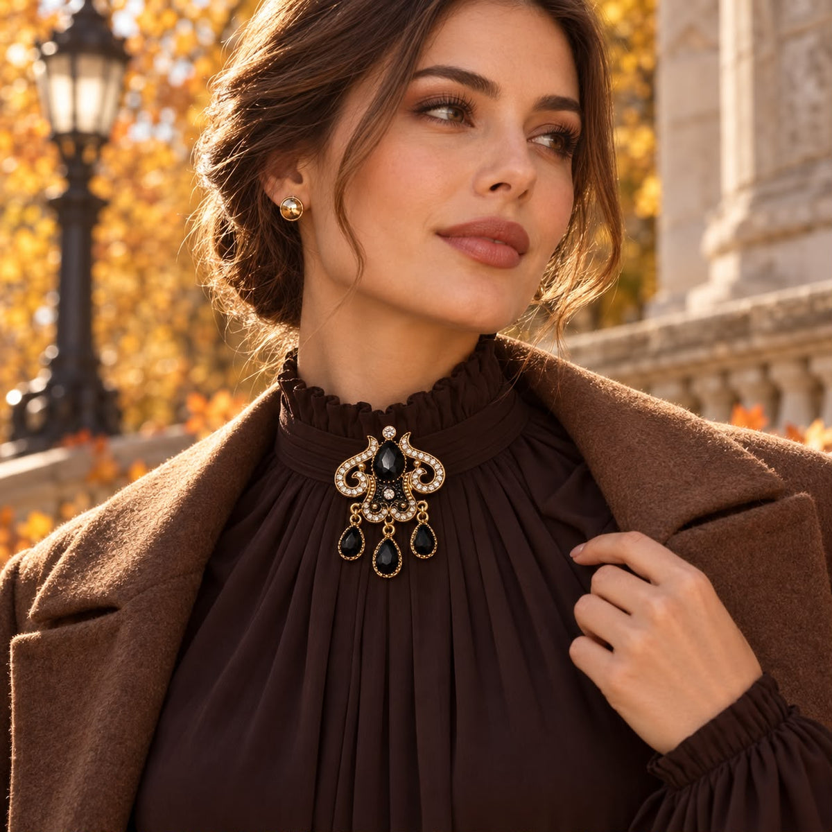 A woman outdoors wearing a dramatic brown high-neck blouse with a large ornate brooch at the ruffled collar, paired with a brown coat. The brooch features gold-toned metalwork, clear rhinestones, and dark blue teardrop stones.